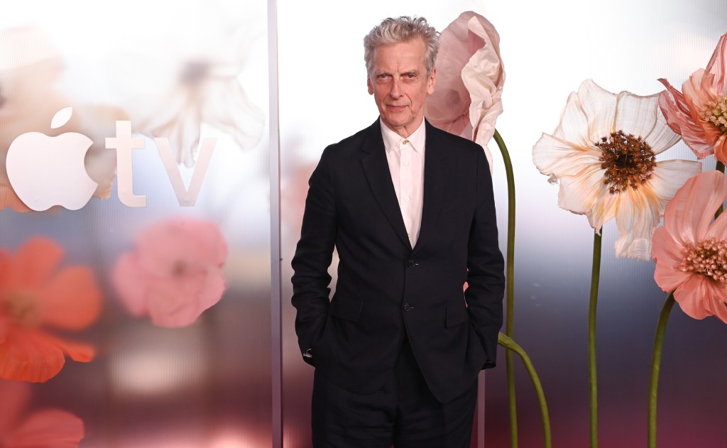 Peter Capaldi in a black suit in front of a poppy wall