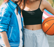 Two faceless girls on a basketball court. One of them is holding a basketball under her arm. There is a banner in the colours of the trans flag behind them.