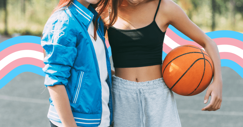 Two faceless girls on a basketball court. One of them is holding a basketball under her arm. There is a banner in the colours of the trans flag behind them.