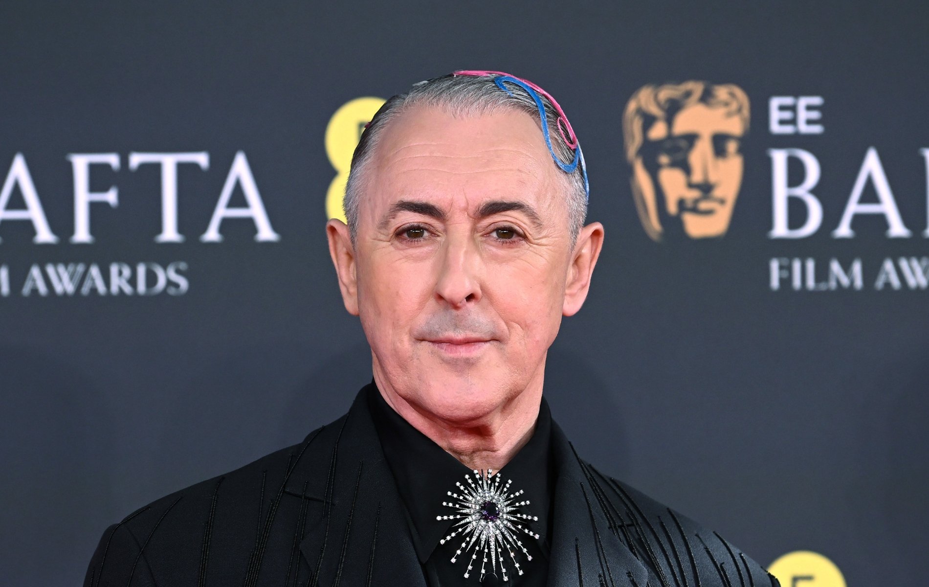 Alan Cumming attends the 2026 EE BAFTA Film Awards at The Royal Festival Hall. He's in a black suit jacket with pink and blue hair accessories. A silver brooche on his tie knot